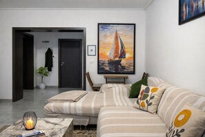 A living room with a sofa, a painting of a sailboat, a potted plant, and a door leading to another room.
