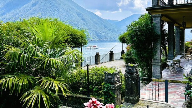 Ein Garten mit rosa Blumen, einer Steinbrücke und Blick auf einen Berg und See.