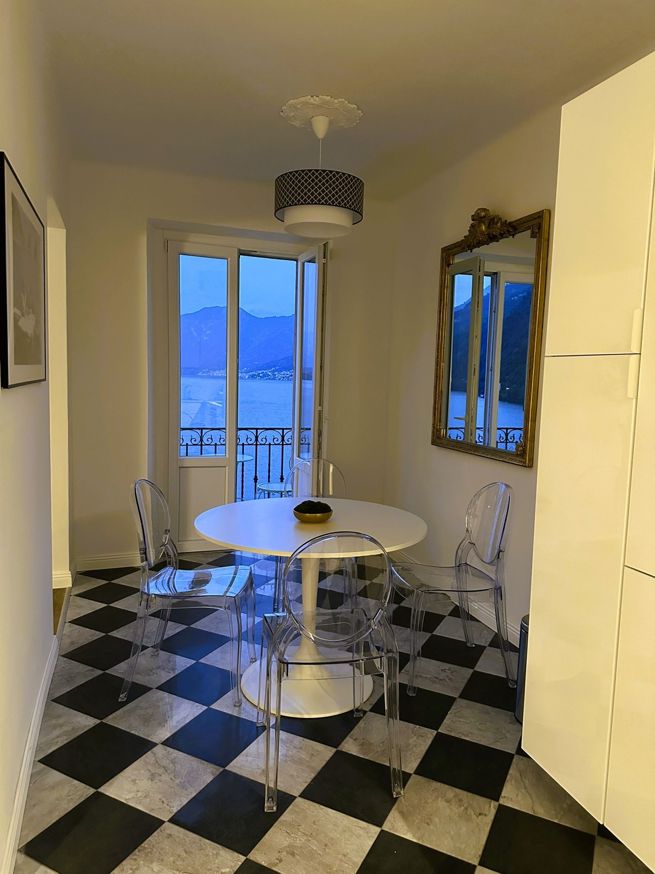 A dining area with a white table and chairs, a checkered floor, a view of mountains through a window, and a ceiling light fixture.