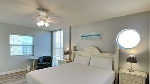 A bedroom with a bed, a ceiling fan, a nightstand with a lamp, a chair, and a window with blinds. - Pelican Isle #513 | Seasonal Beach Service + Resort Amenities (Fort Walton Beach)