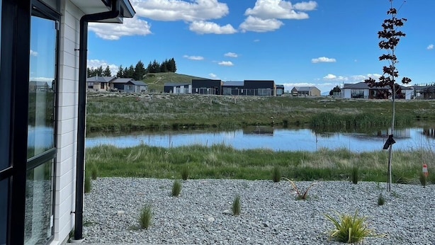 Een modern gebouw met een groot glazen raam, een grindpad met kleine planten, een vijver en een helderblauwe lucht met wolken.
