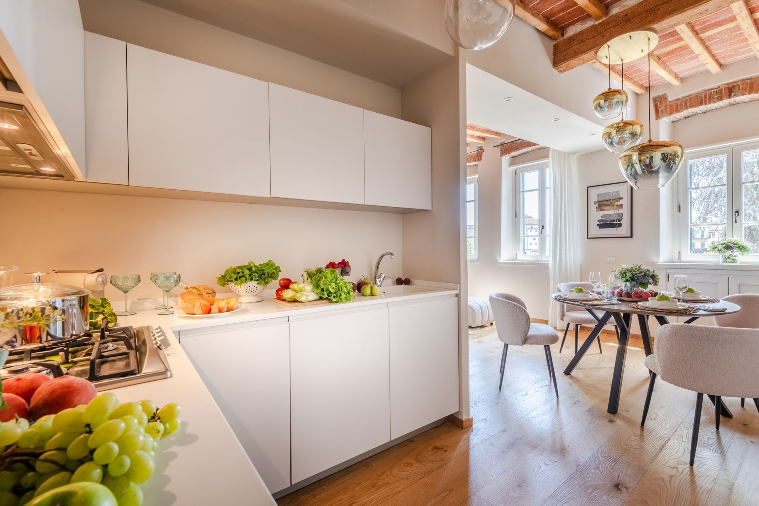 A modern kitchen with white cabinets, a central island with fruits and vegetables, and a dining area with a table and chairs.