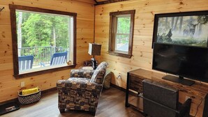A cabin interior with a wooden floor, a patterned armchair, a television, and windows overlooking a forest.