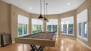 Une table de billard dans une pièce spacieuse, dotée de grandes fenêtres, d’un plancher en bois et de luminaires au plafond.