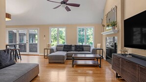 Un salon spacieux avec un canapé d’angle, une table basse et un téléviseur posé sur un meuble en bois. De grandes fenêtres laissent entrer la lumière naturelle et offrent une vue sur la verdure environnante.
