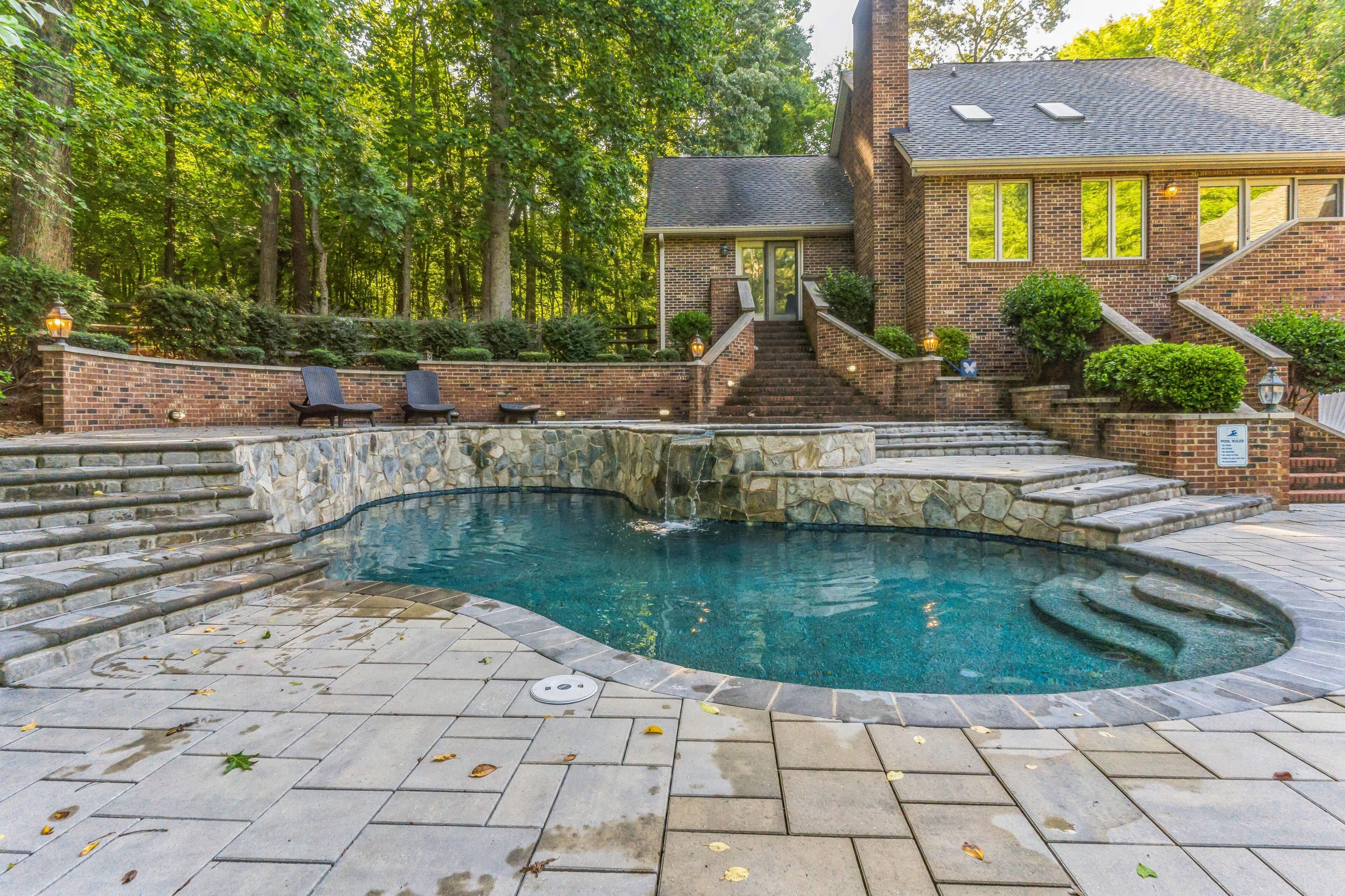 A swimming pool with a stone border, a brick building with a staircase, and a stone pathway.