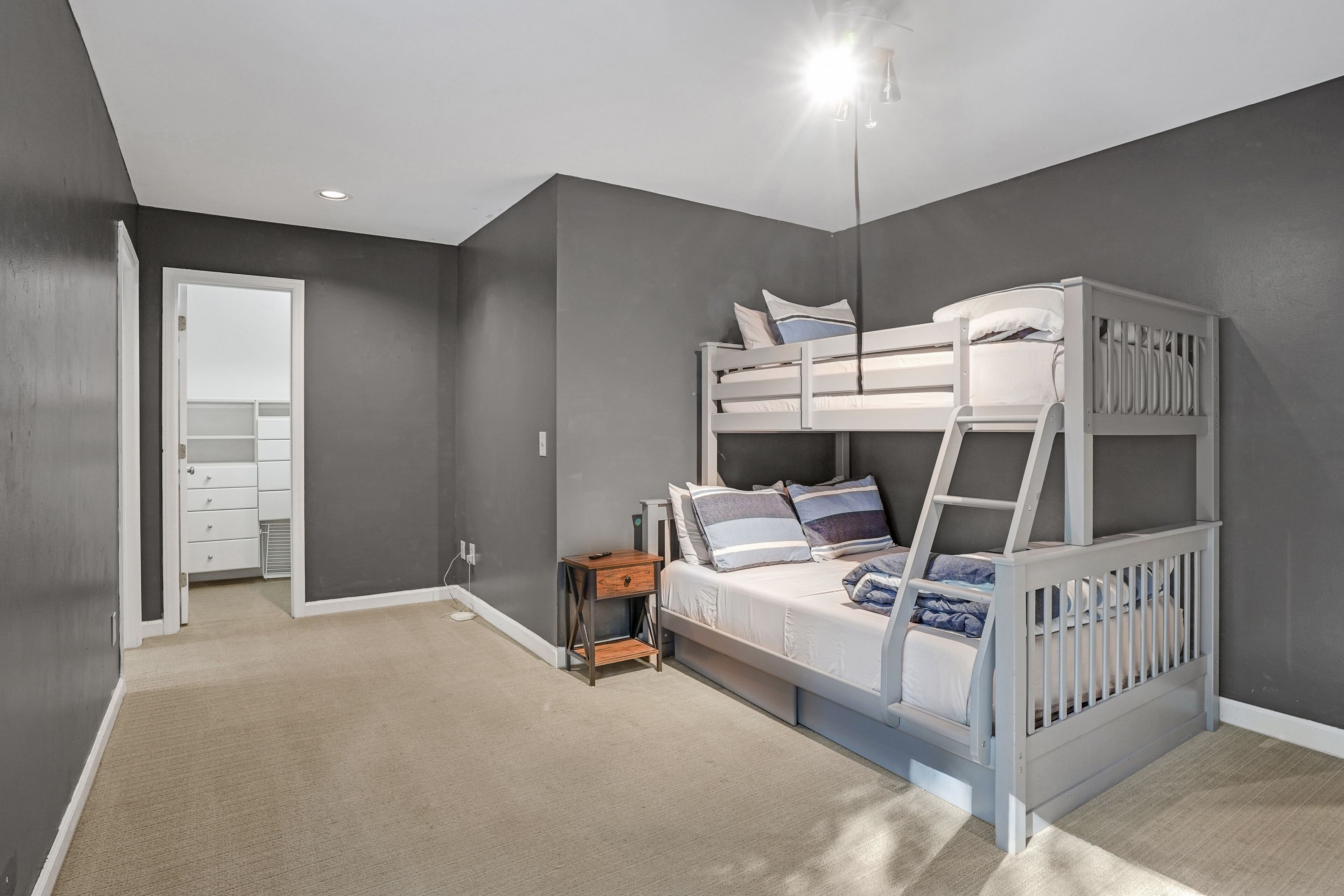 A room with a bunk bed, a small wooden nightstand, a white dresser, and a door leading to another room.