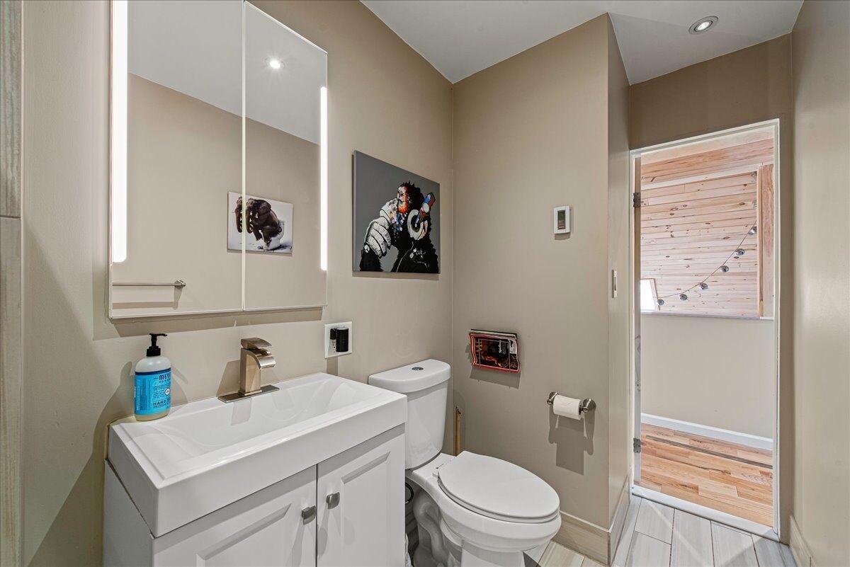 Un bagno moderno con lavandino, specchio e servizi igienici bianchi. Al muro è presente un quadro e una porta in legno dà accesso a una zona esterna.