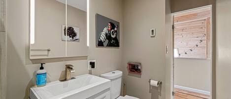 Un bagno moderno con lavandino, specchio e servizi igienici bianchi. Al muro è presente un quadro e una porta in legno dà accesso a una zona esterna.