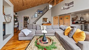 Een ruime woonkamer met een houten plafond, een grijze zithoek, een open haard en een keuken in de achtergrond.