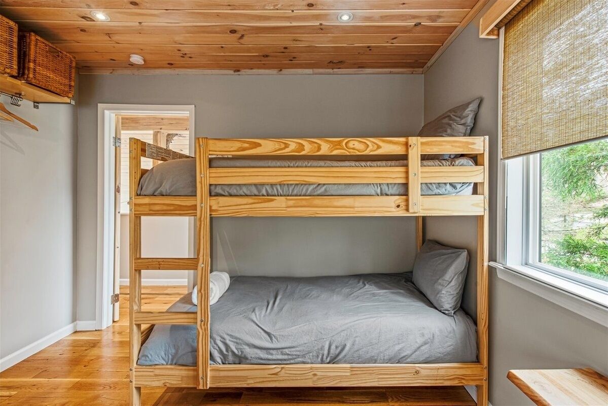 A bunk bed room with a wooden ceiling, grey bedding, and a window with a view of greenery.