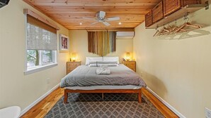 Een slaapkamer met een houten plafond, een bed, een plafondventilator, een raam met zonwering, en een wandairconditioning.