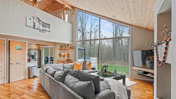 Een ruime woonkamer met een grijze bank, houten vloeren, een televisie en een groot raam met uitzicht op een tuin.