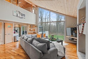 Uma sala de estar espaçosa com um sofá cinzento, piso de madeira, uma televisão e uma grande janela com vista para um jardim.