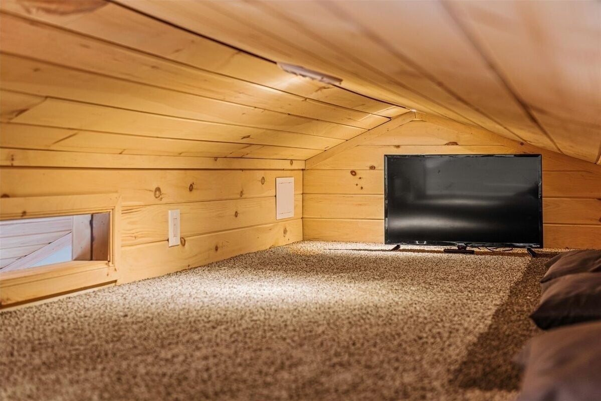 A small attic room with a television, a window, and a carpeted floor.