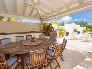 A covered outdoor dining area with a round wooden table and chairs, a ceiling fan, and a view of a swimming pool and greenery in the background. - Ocean View by Bluescape (Belair)