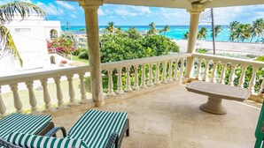 A balcony with two wicker lounge chairs and a stone table, overlooking a beach with palm trees and a clear blue sky. - Ocean View by Bluescape (Belair)