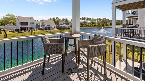 Une terrasse en bois, avec des chaises en osier et une petite table, d’où l’on peut admirer un plan d’eau et des maisons.