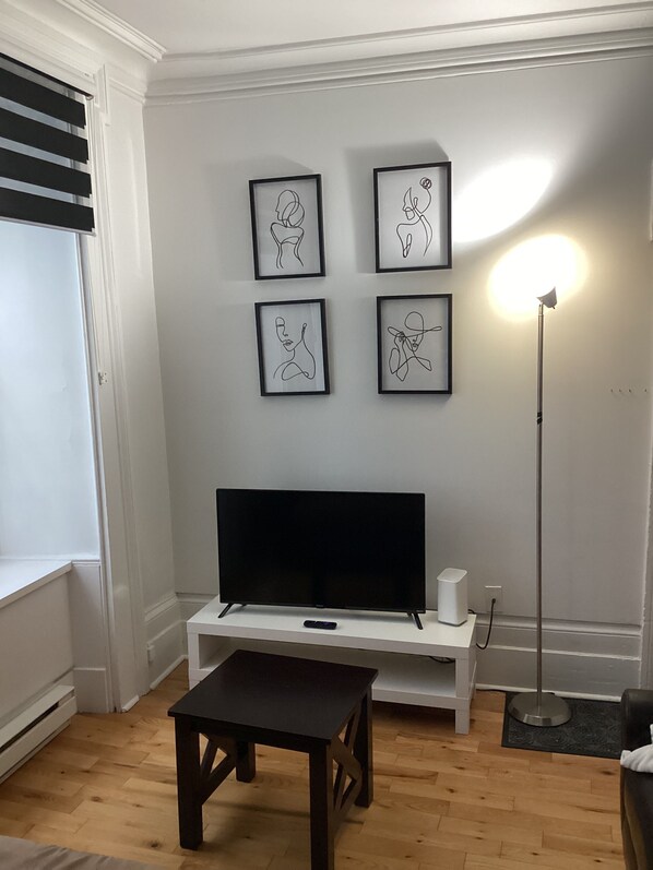 Sala de estar con televisor de pantalla plana, un banco blanco, una lámpara de pie y cuatro cuadros lineales enmarcados en la pared.