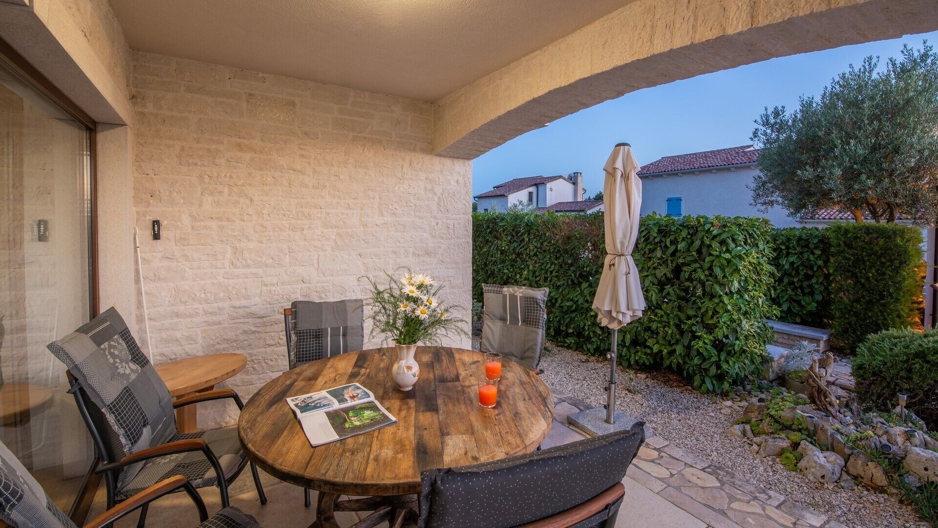 A covered patio with a wooden table, chairs, a vase with flowers, and a glass of orange juice.