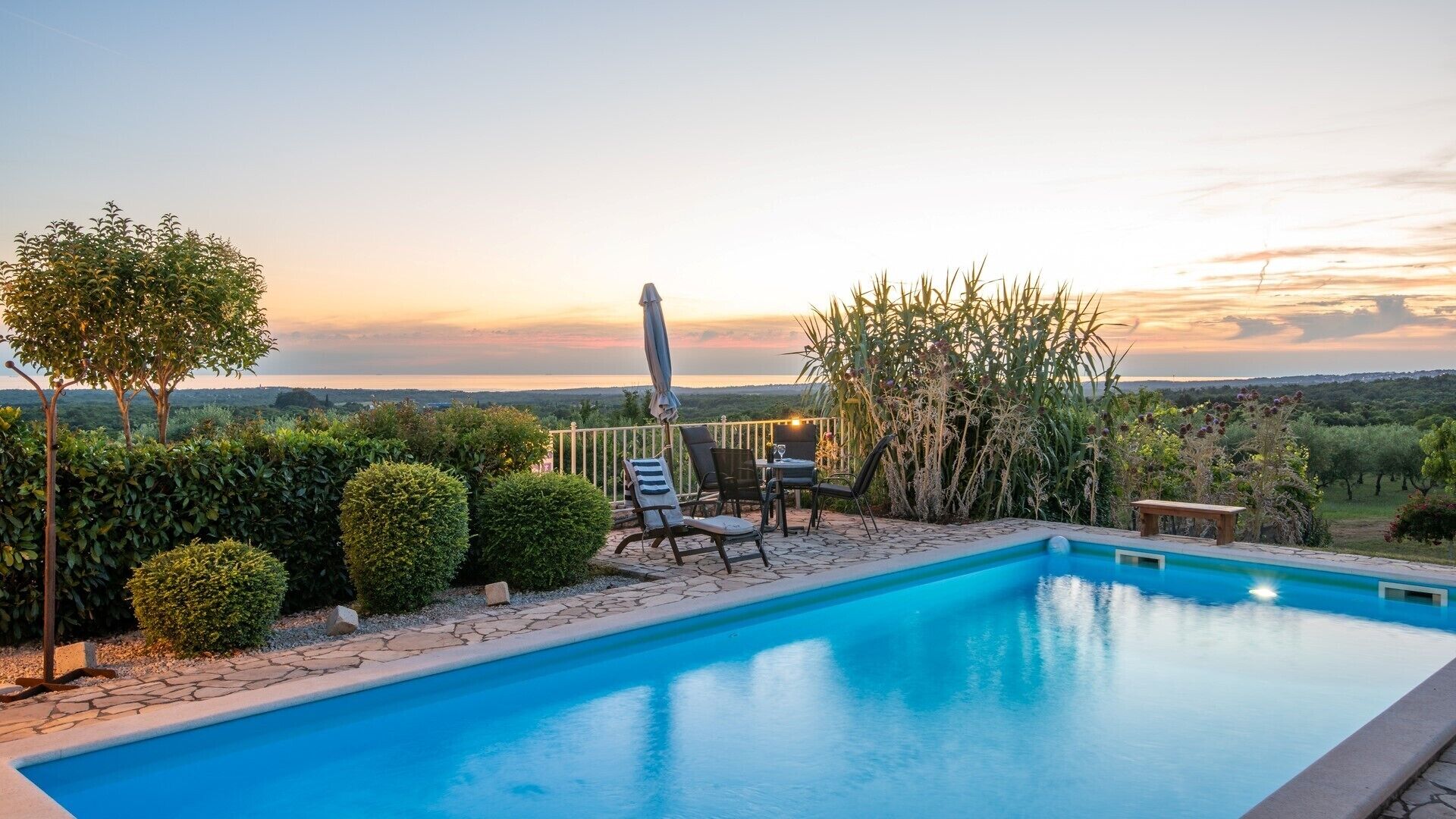 A swimming pool with a view of the sunset, surrounded by neatly trimmed hedges and a patio area with chairs and a table.