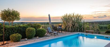A swimming pool with a view of the sunset, surrounded by neatly trimmed hedges and a patio area with chairs and a table.