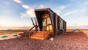 A modern, multi-level wooden structure with a staircase leading to a glass door, situated in a desert-like environment with a clear sky. - Grand Canyon Stargazing Off-Grid Tiny Home (Williams)