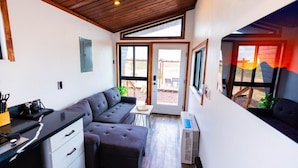 A compact kitchen with a grey sofa, a small dining table, and a window with a view of the outdoors. - Grand Canyon Stargazing Off-Grid Tiny Home (Williams)