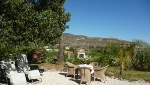 Un patio con mobiliario de mimbre y una mesa, con vistas a un paisaje rural con casas y colinas.