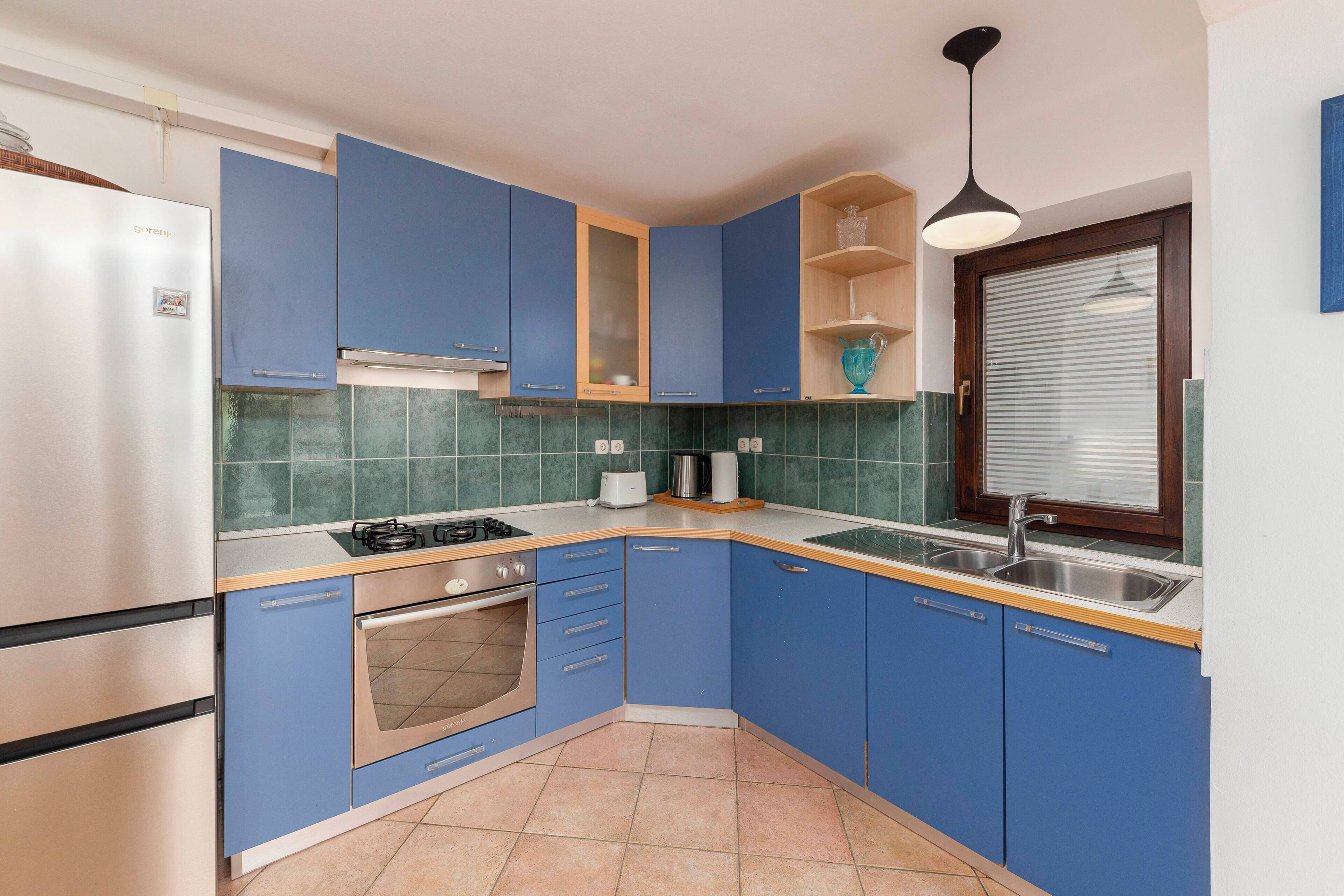 Eine kleine Küche mit blauen Schränken, einem Edelstahl-Kühlschrank, einem eingebauten Ofen und einem Fenster mit Jalousien.