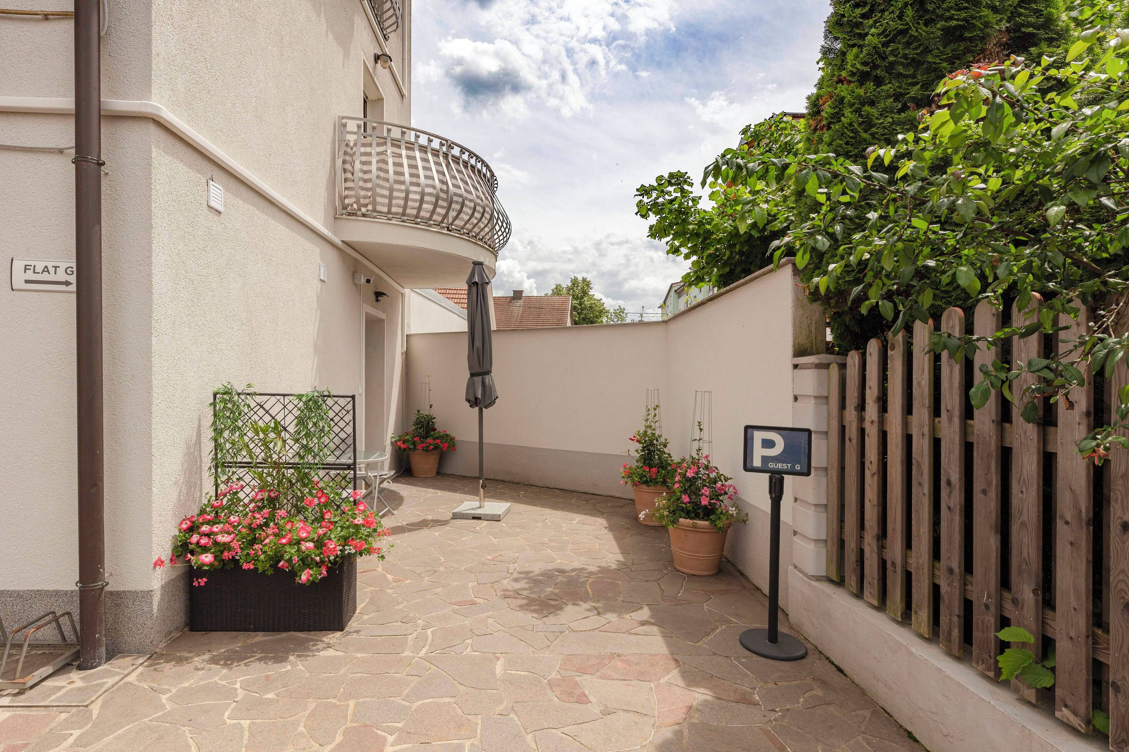 Un patio con zona ajardinada, plantas en macetas, cerca de madera y un cartel que indica zona de estacionamiento para huéspedes.