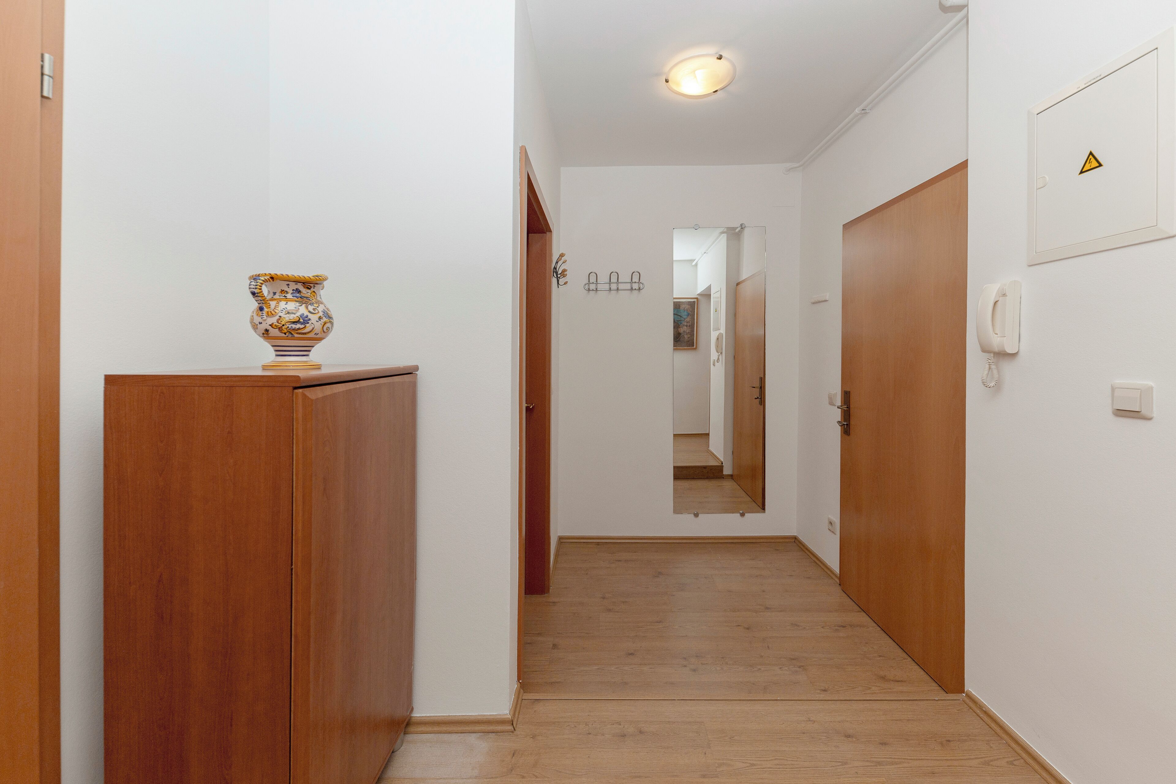 A hallway with wooden flooring, a wooden cabinet, and a vase on top. There are doors on both sides and a light switch on the wall.