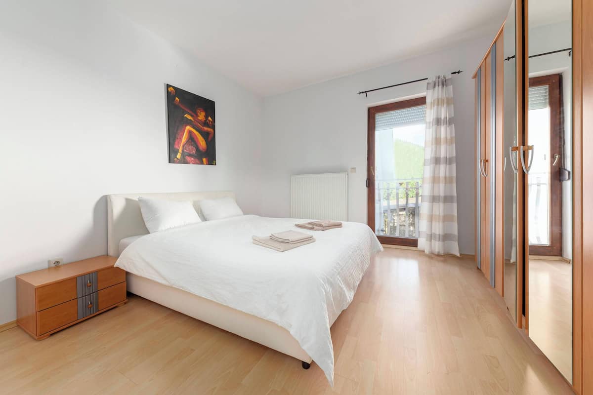 Un dormitorio con cama, mesita de noche, radiador, ventana con cortinas y vistas al exterior.