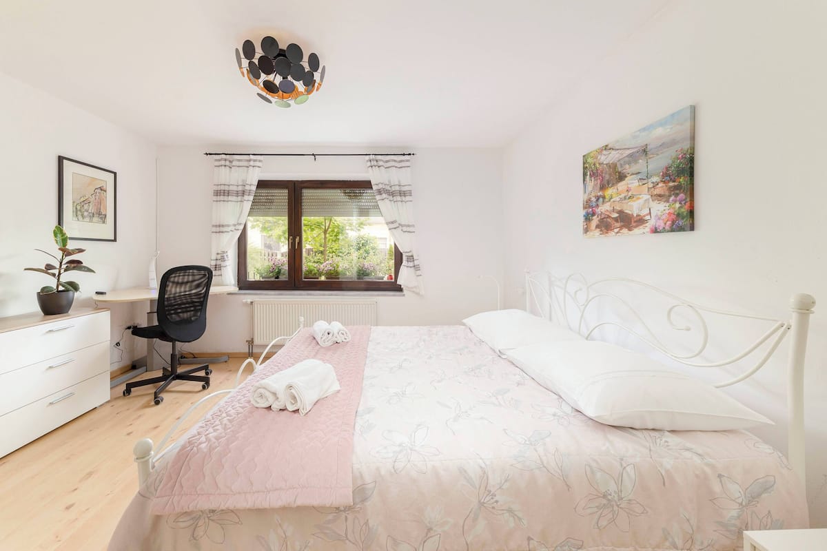 Un dormitorio con cama, escritorio, silla, radiador, ventana con cortinas, una planta y un cuadro en la pared.