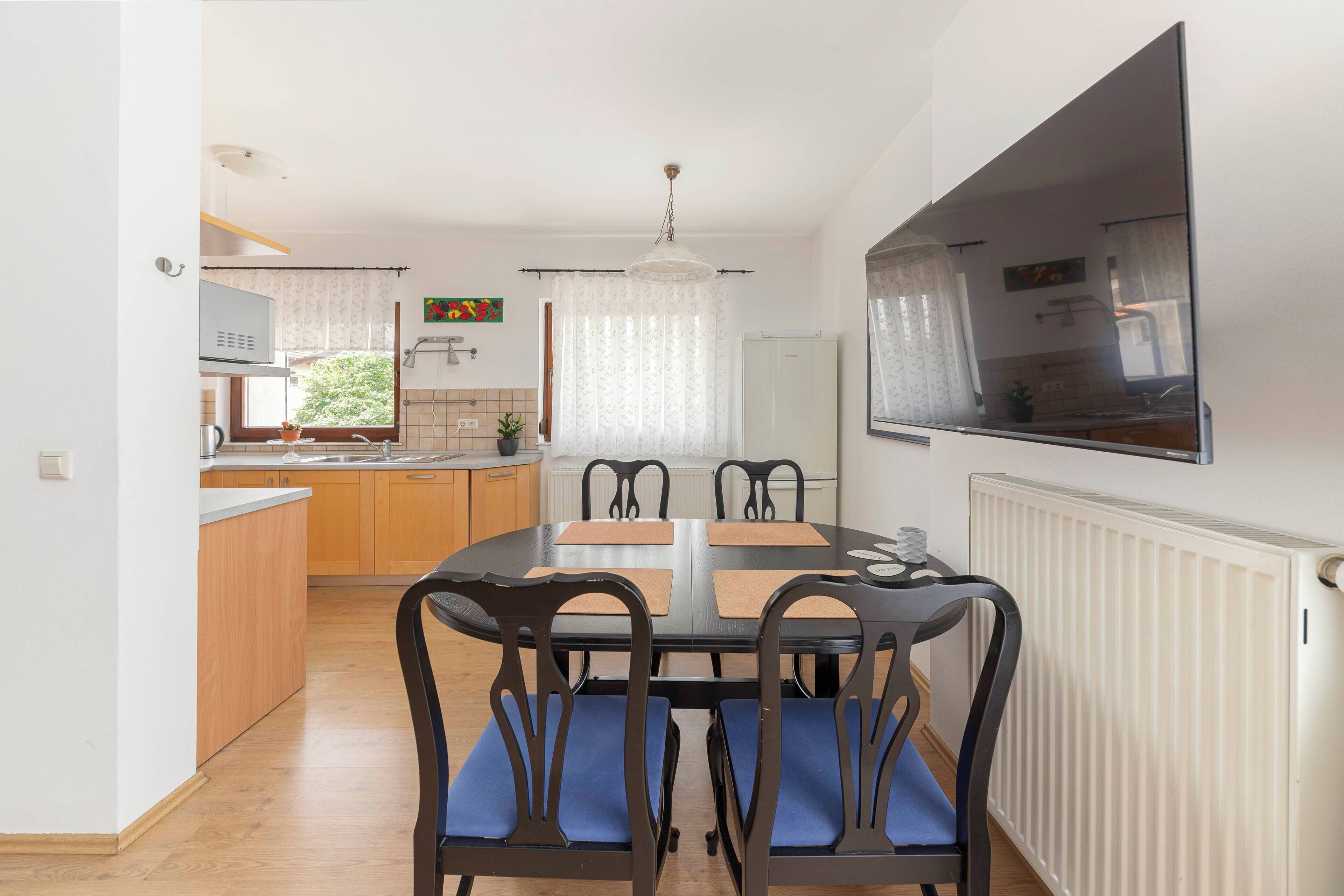 A compact kitchen with wooden cabinets, a dining table with chairs, and a large window with curtains.