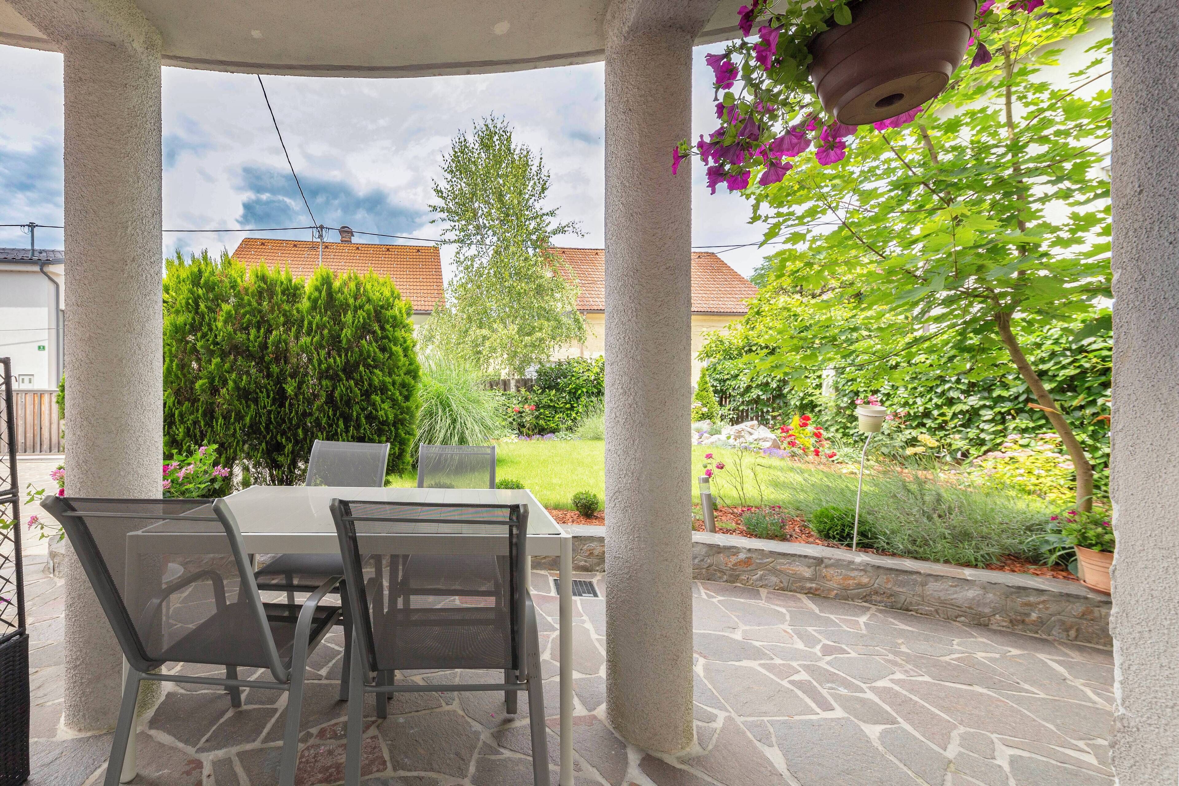 A covered patio area with a stone floor, a table with chairs, and a view of a garden with various plants and a tree.