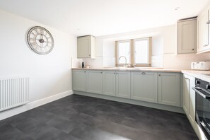 A modern kitchen with grey cabinets, a built-in oven, and a large wall clock. - Host & Stay | Caedmons View (Whitby)