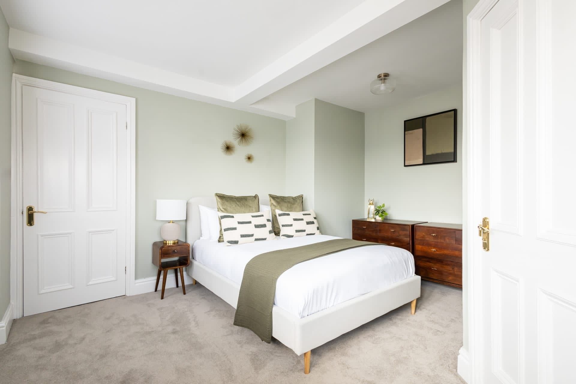 Een slaapkamer met een bed, nachtkastje, lamp, dressoir en een spiegel aan de muur.
