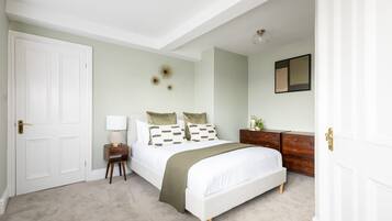 Een slaapkamer met een bed, nachtkastje, lamp, dressoir en een spiegel aan de muur.