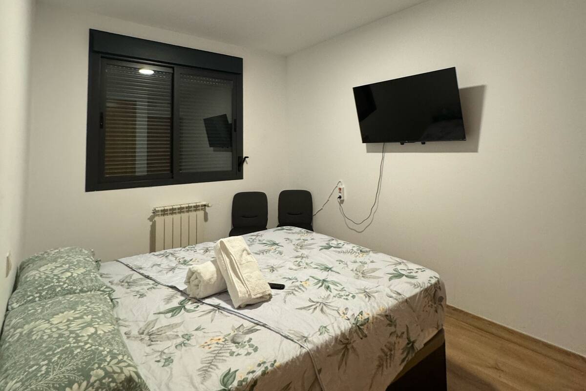Une chambre à coucher avec un lit, une fenêtre avec des stores, un téléviseur fixé au mur et un radiateur.
