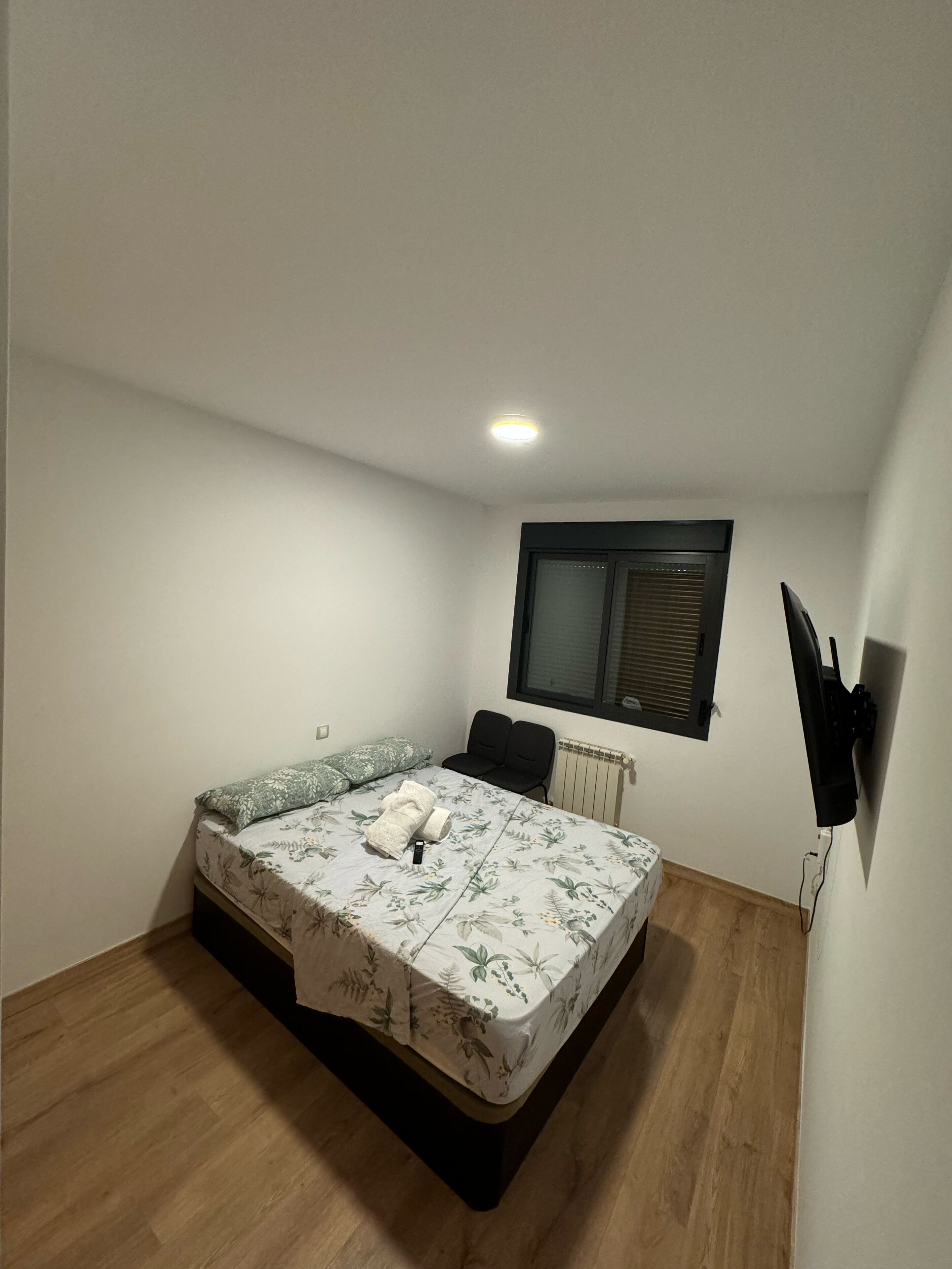 Un dormitorio pequeño y bien iluminado con una cama individual, un radiador, una ventana con persianas y un televisor montado en la pared.