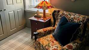 A room with a floral-patterned sofa, a small wooden nightstand, a lamp, a mirror, and a window with blinds. - Walk to Main St! Spacious 2BR Bi-Level • Sleeps 8 (Philadelphia)