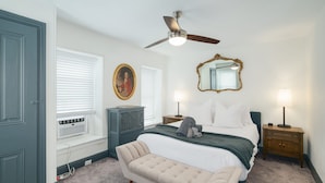 A bedroom with a bed, bedside lamps, a ceiling fan, a window with blinds, a radiator, a bench, and a mirror on the wall. - Walk to Main St! Spacious 2BR Bi-Level • Sleeps 8 (Philadelphia)