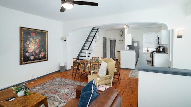 A spacious living room with a brown leather couch, a wooden coffee table, and a painting on the wall. A dining area with a wooden table and chairs is visible in the background. - Walk to Main St! Spacious 2BR Bi-Level • Sleeps 8 (Philadelphia)