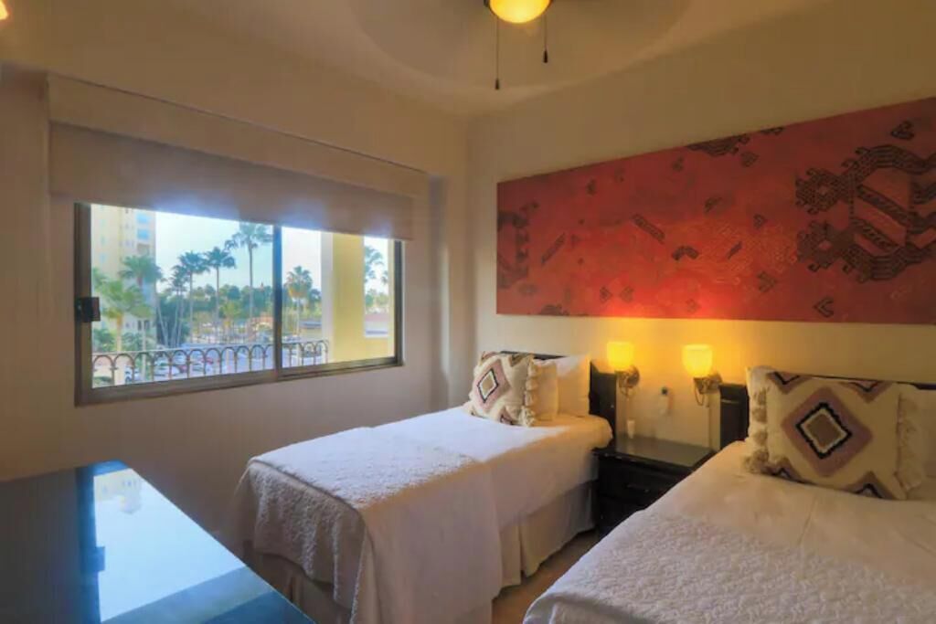 Habitación de hotel con dos camas, un ventanal con vistas a palmeras y una pared con diseño rojo.