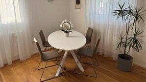 A dining area with a round table, four chairs, a potted plant, a clock on the wall, and sheer curtains.