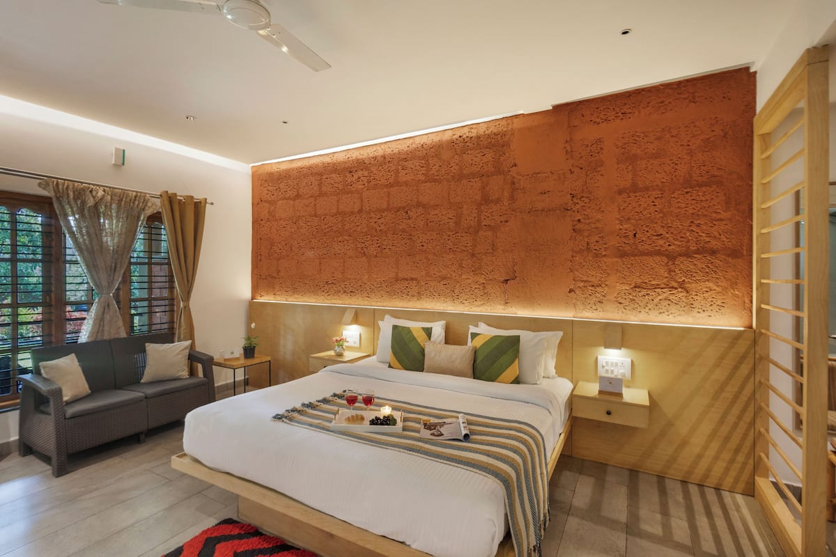 A modern hotel room with a large bed, a sofa, a nightstand with a lamp, and a view of the outdoors through the window.