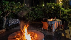 An outdoor patio area with a circular fire feature, a table with chairs, and a table with a floral tablecloth. - Avocado Cottages by Alaya stays (Somvarpet)