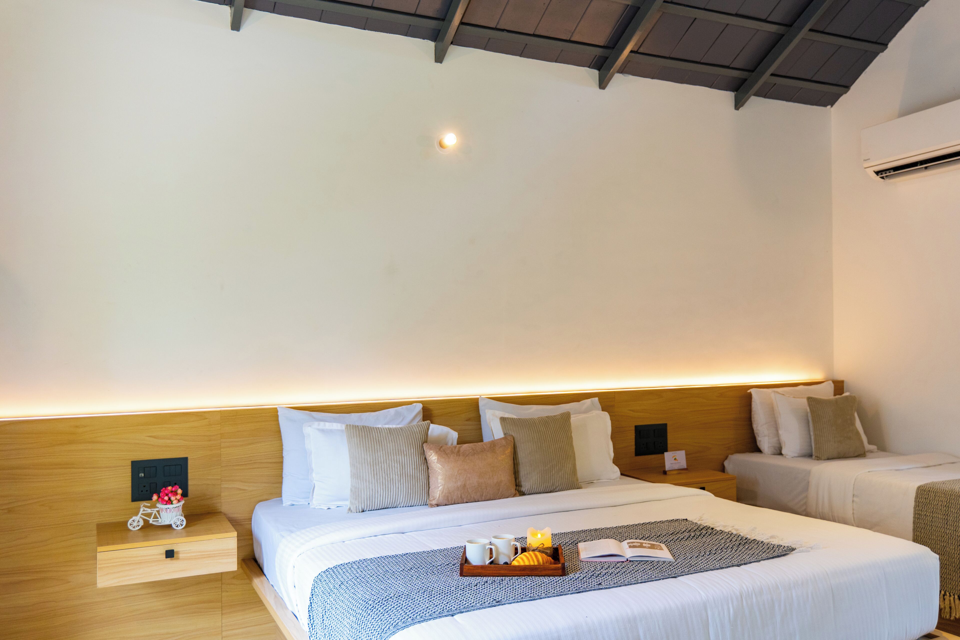 A hotel room with two beds, a bedside table with a lamp, a small shelf with decorative items, and a ceiling light.
