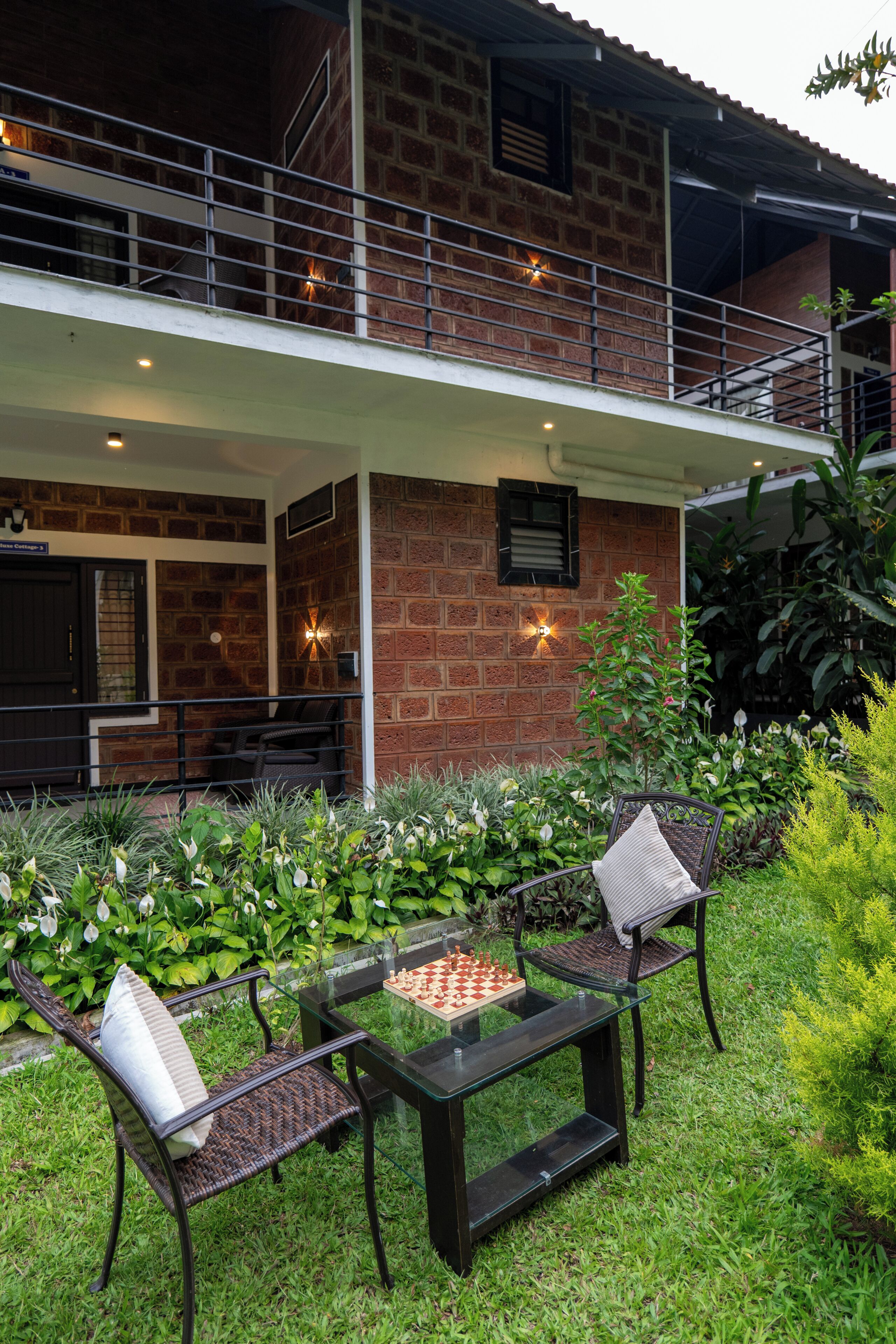 A two-story building with a brick exterior, a balcony with metal railings, and a chess table in the garden area.